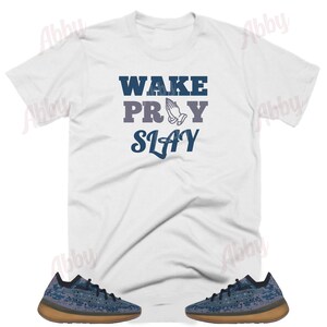 Puede incluir: Camiseta blanca con el texto "WAKE PRAY SLAY" en letras azules. La palabra "PRAY" tiene un gr&aacute;fico de manos en oraci&oacute;n. Un par de zapatillas azules y marrones se encuentran en las esquinas inferiores de la imagen.