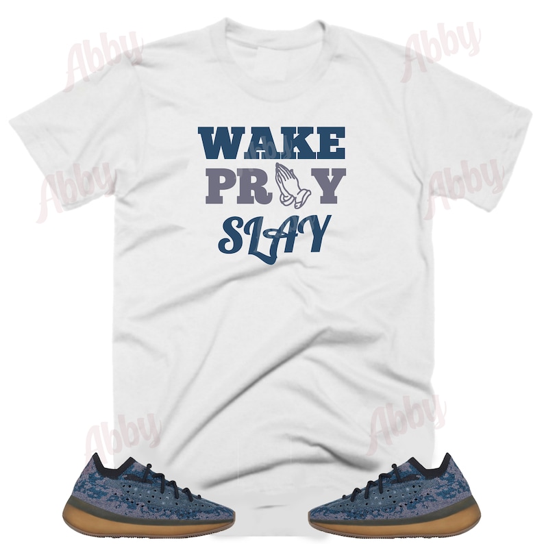 Puede incluir: Camiseta blanca con el texto "WAKE PRAY SLAY" en letras azules. La palabra "PRAY" tiene un gr&aacute;fico de manos en oraci&oacute;n. Un par de zapatillas azules y marrones se encuentran en las esquinas inferiores de la imagen.