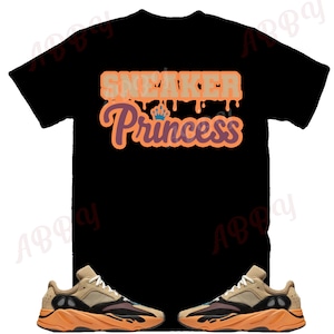May include: Black t-shirt with the text "Sneaker Princess" in orange and brown lettering. The shirt is paired with two tan and orange sneakers.