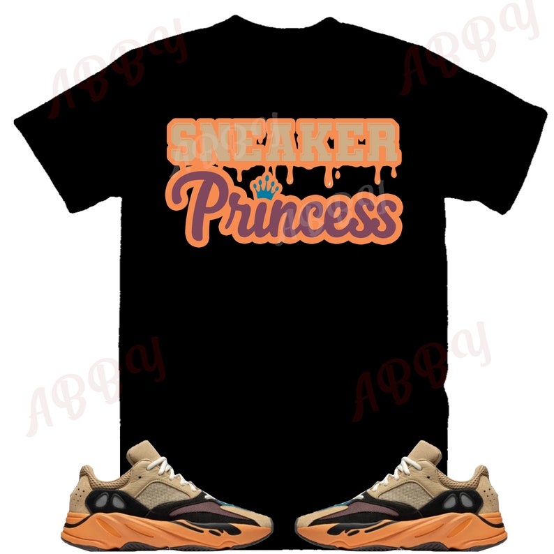 May include: Black t-shirt with the text "Sneaker Princess" in orange and brown lettering. The shirt is paired with two tan and orange sneakers.