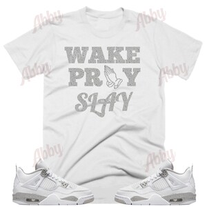 May include: White t-shirt with the text "WAKE PRAY SLAY" in gray glitter lettering. A pair of white and gray sneakers with a gray speckled design.