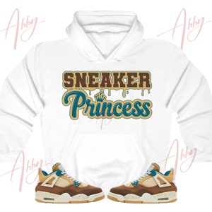 May include: White hoodie with the text "Sneaker Princess" in brown, teal, and gold lettering. The hoodie is paired with a pair of brown, teal, and white sneakers.