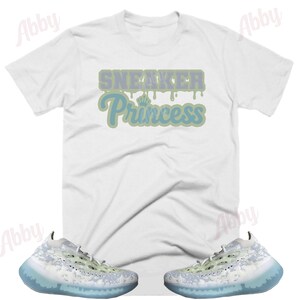 May include: White t-shirt with the text "Sneaker Princess" in blue and green lettering. A pair of white and light green sneakers with a blue translucent sole.