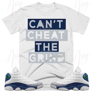 May include: White t-shirt with the text "Can't Cheat The Grind" in navy blue and gray lettering. A pair of white, gray, and navy blue sneakers are shown on either side of the t-shirt.