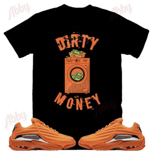 Peut inclure: Un t-shirt noir avec un graphique orange d'une machine &agrave; laver contenant de l'argent. Le texte "DIRTY MONEY" est imprim&eacute; sous le graphique. Une paire de baskets orange est &eacute;galement repr&eacute;sent&eacute;e.