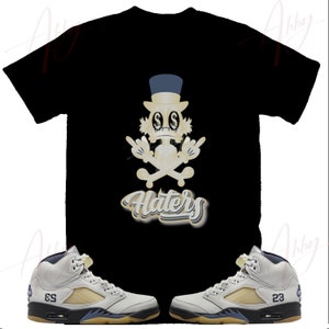 May include: A black t-shirt with a cartoon character wearing a top hat and holding a skull and crossbones. The text "Haters" is written below the character. The t-shirt is paired with a pair of grey and white sneakers with blue accents.