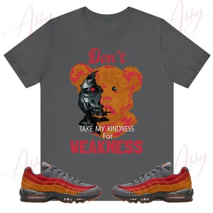 May include: Dark gray t-shirt featuring a teddy bear design with a cyborg face and the text "Don't Take My Kindness For Weakness" in red. Below the shirt are orange, red, and gray sneakers.
