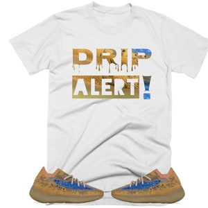 May include: White t-shirt with the text "DRIP ALERT!" in a dripping font with a desert-like pattern. The shirt is paired with a pair of tan and blue sneakers.