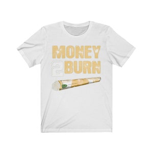 Puede incluir: Camiseta blanca con el texto "MONEY 2 BURN" en letras doradas y una imagen de dibujos animados de un porro enrollado.