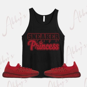 Peut inclure: D&eacute;bardeur noir avec le texte "SNEAKER Princess" en rouge. Deux baskets rouges sont plac&eacute;es de chaque c&ocirc;t&eacute; du d&eacute;bardeur. Les baskets ont un design textur&eacute;.