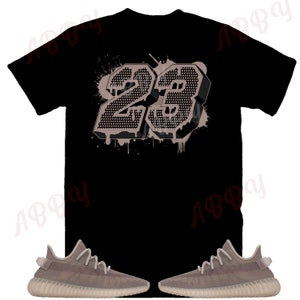 Op de afbeelding: Een zwart T-shirt met een groot, grijs, 3D-nummer "23" gedrukt op de achterkant. Het nummer heeft een verfspatten-effect. Het T-shirt wordt gecombineerd met een paar grijze sneakers.