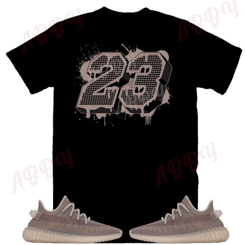 Op de afbeelding: Een zwart T-shirt met een groot, grijs, 3D-nummer "23" gedrukt op de achterkant. Het nummer heeft een verfspatten-effect. Het T-shirt wordt gecombineerd met een paar grijze sneakers.