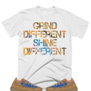 May include: White t-shirt with the text "GRIND DIFFERENT SHINE DIFFERENT" in a layered, textured font. The shirt is paired with a pair of tan and blue trainers.