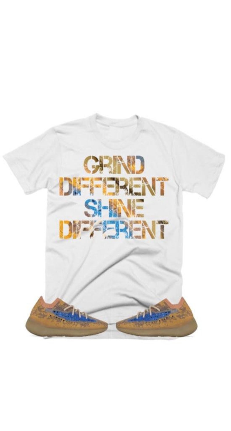 May include: White t-shirt with the text "GRIND DIFFERENT SHINE DIFFERENT" in a layered, textured font. The shirt is paired with a pair of tan and blue trainers.