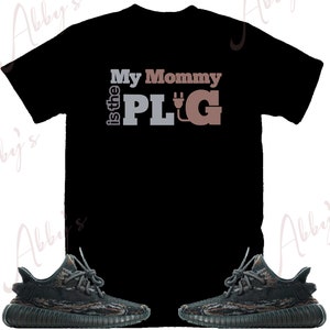 May include: Black t-shirt with the text "My Mommy is the PLUG" in brown and gray lettering. The shirt is paired with a pair of black and green camouflage sneakers.