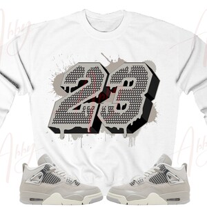 May include: White long-sleeve sweatshirt with a gray and black number "23" graphic. The number is in a 3D style with a dripping paint effect. Two pairs of gray and white sneakers are shown below the sweatshirt.