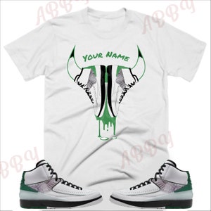 May include: White t-shirt with a green and black bull head graphic featuring a pair of white, black, and green sneakers. The text "YOUR NAME" is printed on the t-shirt. A pair of white, black, and green sneakers are also shown.