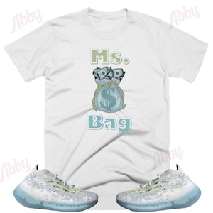 Puede incluir: Camiseta blanca con un gr&aacute;fico de una bolsa de dinero con el texto "Ms. Bag". Un par de zapatillas blancas y azul claro con un patr&oacute;n de camuflaje.