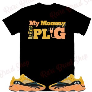 Puede incluir: Camiseta negra con el texto "My Mommy is the PLUG" en naranja y amarillo. Dos pares de zapatillas amarillas, negras y verdes con suelas naranjas.
