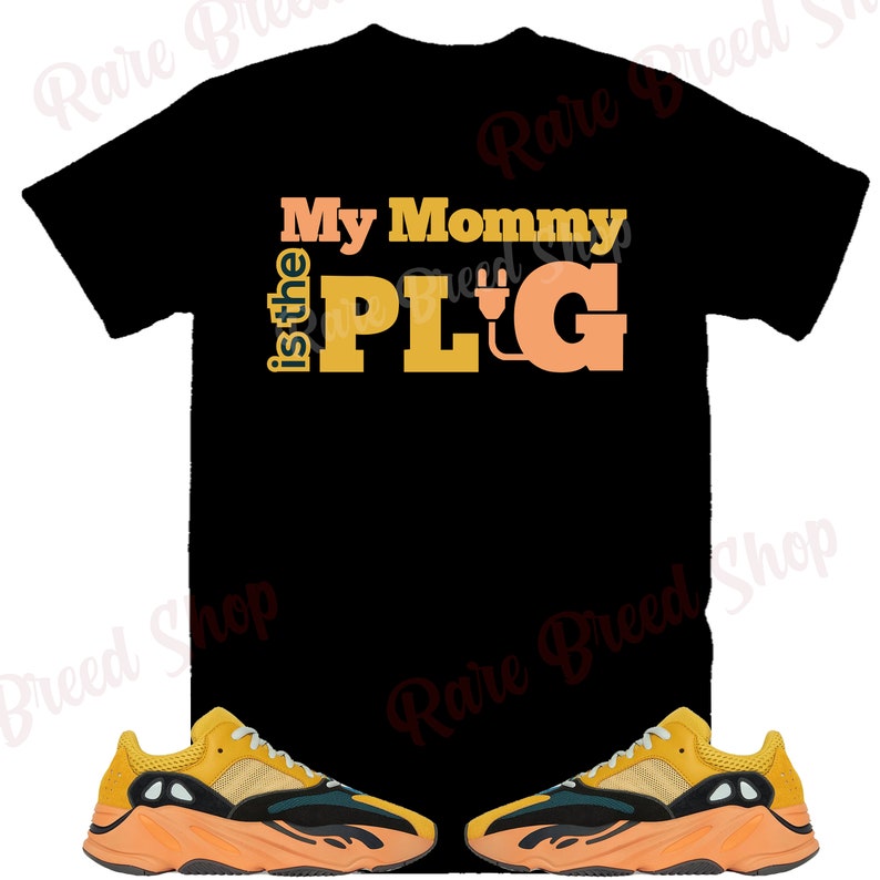 Puede incluir: Camiseta negra con el texto "My Mommy is the PLUG" en naranja y amarillo. Dos pares de zapatillas amarillas, negras y verdes con suelas naranjas.