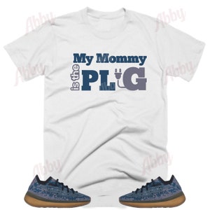 May include: White t-shirt with the text "My Mommy is the PLUG" in blue and gray lettering with an electric plug graphic. The shirt is paired with a pair of blue and brown sneakers.