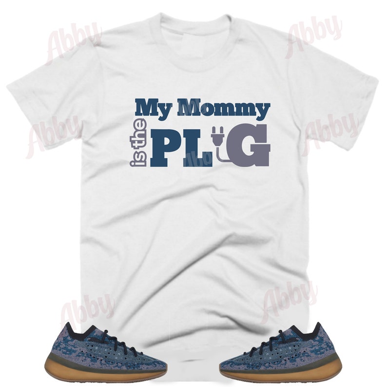 May include: White t-shirt with the text "My Mommy is the PLUG" in blue and gray lettering with an electric plug graphic. The shirt is paired with a pair of blue and brown sneakers.