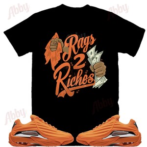 以下が含まれることがあります： オレンジ色のグラフィックがプリントされた黒いTシャツ。グラフィックには「Rags 2 Riches」の文字と、赤い布を持っている手と、お金を持っている手が描かれている。オレンジ色のスニーカーには銀色のアクセントが施されている。