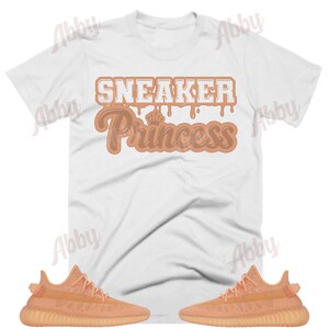 May include: A white t-shirt with the text "Sneaker Princess" in brown dripping letters. The shirt is paired with two pairs of light orange sneakers.
