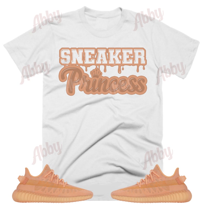 May include: A white t-shirt with the text "Sneaker Princess" in brown dripping letters. The shirt is paired with two pairs of light orange sneakers.