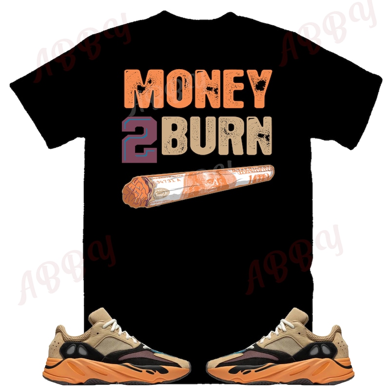 Op de afbeelding: Zwart T-shirt met de tekst "MONEY 2 BURN" in oranje en paars. Een opgerolde dollarbiljet is afgebeeld onder de tekst. Twee paar beige en oranje sneakers bevinden zich aan beide kanten van het T-shirt.