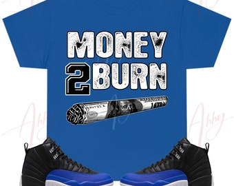 May include: A blue t-shirt with the text "MONEY 2 BURN" in white with a black and white image of a rolled-up dollar bill. The shirt is paired with black and blue sneakers.