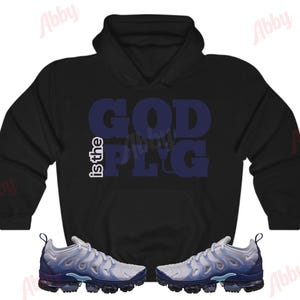 May include: Black hooded sweatshirt with the text "GOD is the PLUG" in blue. The sweatshirt is paired with a pair of white and blue sneakers.