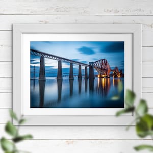 May include: A photograph of the Forth Rail Bridge in Scotland at dusk. The bridge is a steel structure with a red colour. The bridge is reflected in the water below. The sky is a light blue colour with some clouds.