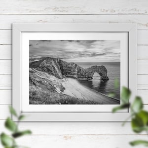 May include: A black and white photograph of a natural stone archway on a rocky coastline. The archway is formed by the erosion of the cliff face, creating a natural bridge over the ocean. The beach below the archway is covered in sand and pebbles.