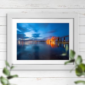 May include: A photograph of a bridge over a body of water at dusk. The bridge is lit up with orange lights, and the water reflects the lights. The sky is a deep blue with clouds.