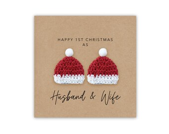 May include: A Christmas card with a brown background features two red and white crocheted Santa hats with white pom-poms. The text reads "HAPPY 1ST CHRISTMAS AS Husband & Wife."