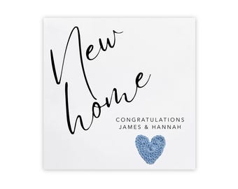 May include: A white card with the words "New Home" in black script. Below, it reads "CONGRATULATIONS JAMES & HANNAH." A blue crocheted heart is at the bottom. A simple, elegant design for a housewarming or congratulatory gift.