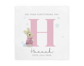 May include: A pink and white card with a large pink letter H and a crocheted bunny in a pink dress. The card says "On your christening day" and "Hannah" with the date "25th July 2024".