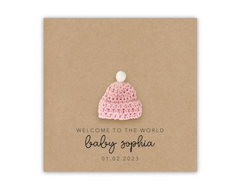 May include: A brown card with a pink crocheted hat with a white pom-pom on top. The card says "Welcome to the world baby sophia 01.02.2023"