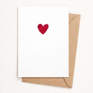 Peut inclure: Une carte de vœux blanche avec un cœur rouge crocheté au centre, posée sur une enveloppe marron. La carte et l'enveloppe sont posées sur un fond neutre, créant un design simple et élégant.