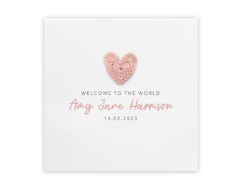 May include: A white card with a crocheted pink heart at the top. The text "WELCOME TO THE WORLD" is above the name "Amy Jane Harrison" and the date "13.02.2023" in a soft pink font. The card is a baby announcement.