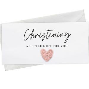 May include: A white card with the word "Christening" in elegant script, accompanied by the phrase "A LITTLE GIFT FOR YOU." A small, pink crocheted heart is affixed to the card. The card is placed on top of a white envelope.