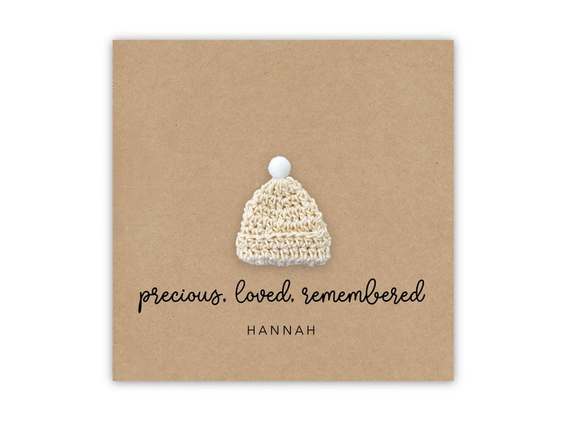 Puede incluir: Un peque&ntilde;o gorro blanco de ganchillo con un pomp&oacute;n blanco en la parte superior. El gorro est&aacute; sobre un fondo marr&oacute;n con el texto "precious. loved. remembered" y el nombre "HANNAH" debajo.