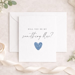 Könnte beinhalten: Eine weiße Karte mit dem Text "WILL YOU BE MY something blue?" und einem blauen gehäkelten Herzen. Die Karte liegt auf einem weißen Umschlag, mit weißen Blumen und einem Band im Hintergrund.