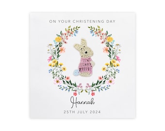 May include: A white card with a floral wreath surrounding a crocheted bunny in pink and white. The card says "On your christening day" and "Hannah" with the date "25th July 2024".