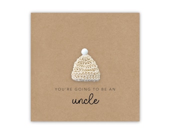 Peut inclure: Une carte marron avec un petit bonnet de bébé en crochet blanc avec un pompon blanc sur le dessus. La carte dit "You're going to be an uncle" en texte noir.