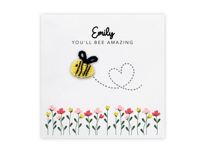 May include: A white card with the name "Emily" and the text "You'll bee amazing" in black lettering. A yellow crocheted bee with black stripes and wings is flying towards a gray dotted heart. A row of green stems with pink and yellow flowers are at the bottom of the card.