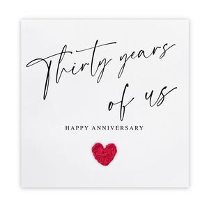 Może przedstawiać: Biała kartka rocznicowa z napisem "Thirty years of us" elegancką czcionką w kolorze czarnym. Poniżej znajduje się napis "Happy Anniversary" i mały, czerwony, sercowaty detal.