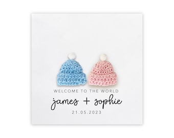 May include: A white card with two crocheted hats, one blue and one pink, with white pom-poms. The text "Welcome to the world james + sophie 21.05.2023" is printed below the hats.