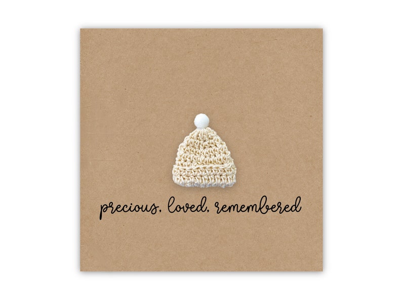 Puede incluir: Una tarjeta marr&oacute;n con un peque&ntilde;o gorro blanco de ganchillo y el texto "precious. loved. remembered."